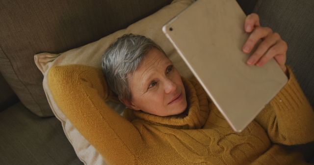 Senior woman lies on a couch holding a tablet while relaxing. She wears a yellow sweater, focusing on the screen. Useful for depicting senior lifestyle, technology use among elderly, or relaxation at home themes.