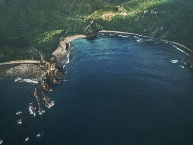 Aerial view capturing a secluded coastline with rocky formations meeting deep blue ocean water and lush green hills in the background. Ideal for use in travel and tourism promotions, nature and adventure blogs, environmental conservation reports, and decorative wall art for coastal-themed interiors.