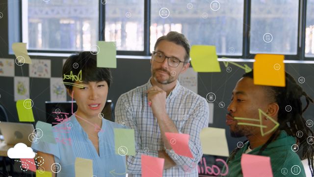 Diverse group of colleagues brainstorming and planning on a glass wall with sticky notes. Digital icons overlay suggests high-tech and interconnected environment. Suitable for depicting teamwork, creative planning, collaborative strategy sessions in a modern office setting with an emphasis on innovation and networking.