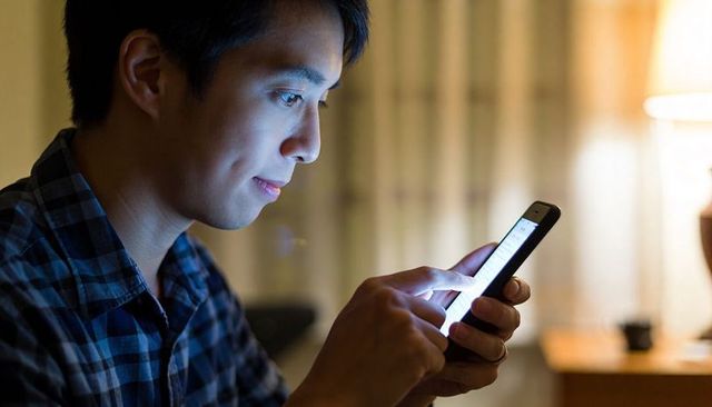 Young asian man scrolling smartphone in dim bedroom at night with blue screen glow, plaid shirt