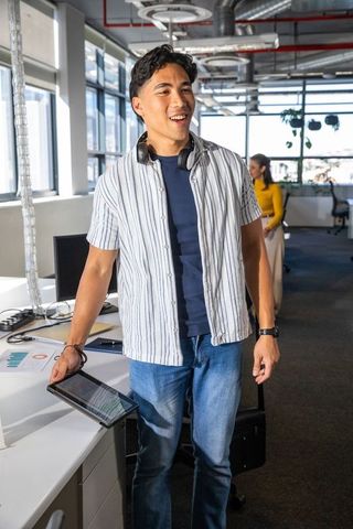 Young Asian man smiling while holding tablet in bright collaborative open-plan office space