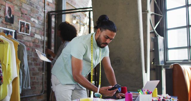 Fashion designer uses smartphone in creative studio with measuring tape around neck, surrounded by fabrics, laptop and colorful threads. Perfect for illustrating modern workspaces in the fashion industry, young professionals in creative fields, and the technology-driven fashion design process.