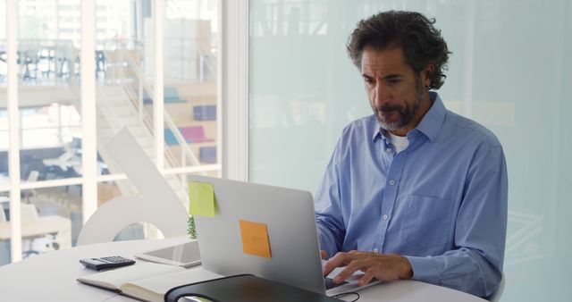 Mature businessman wearing blue shirt working on a laptop in a modern office. The workspace appears organized with sticky notes on the laptop, documents on the table, and bright, natural light coming through the windows. Ideal for illustrating concepts of remote work, business activities, productivity, and professional communication.