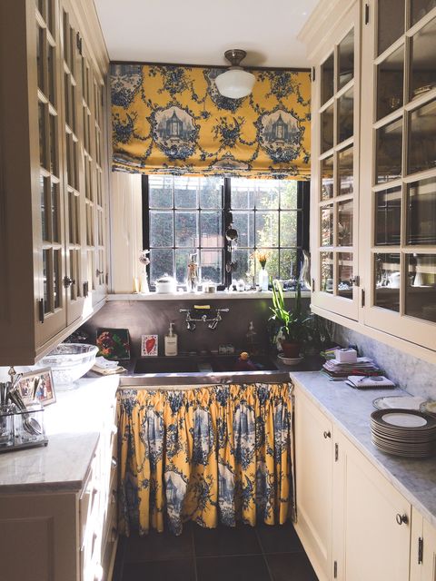 Charming vintage style kitchen decorated with patterned yellow and blue curtains and drapes. Natural light streaming through large windows illuminates the space. The design includes classic glass-front cabinets and a double-bowl sink. Ideal for use in home decor, interior design magazines, and kitchen renovation ideas.