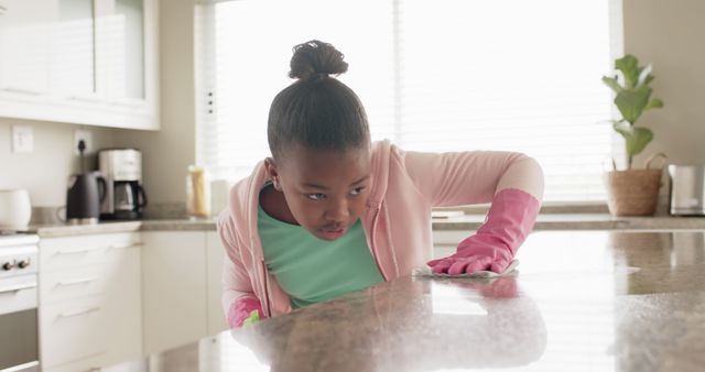Young girl wearing pink gloves cleaning kitchen countertop with determined expression. Proper sanitation and cleanliness concept. Useful for articles on teaching kids household chores, encouraging responsibility, home cleanliness, and hygiene practices.