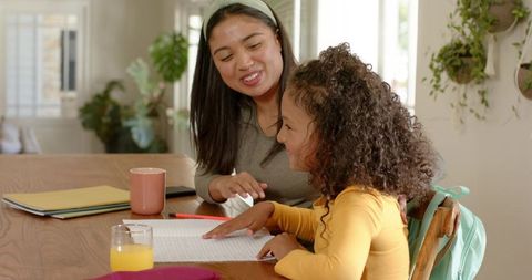 Mother tutoring daughter at home helping with homework at sunny dining table
