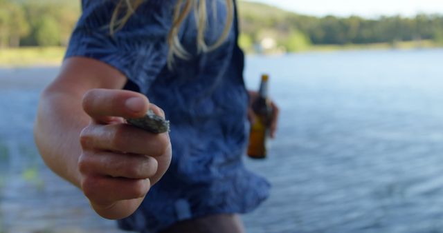 Scene shows person by the lake skipping stone while holding beer bottle. Ideal for themes of summer relaxation, outdoor activities, nature enjoyment, and leisure time. Great for lifestyle blogs, outdoor activity promotions, and social media posts focusing on summer fun.
