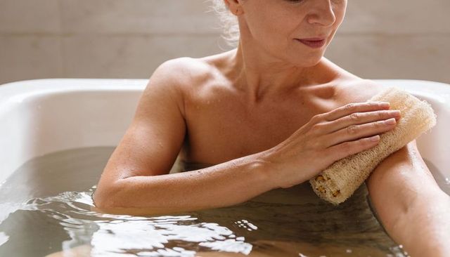 Mature woman bathing and scrubbing shoulder with loofah in relaxing tub for self care