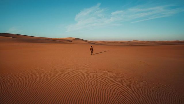 Solitary man walking across rippled sand dunes casting long shadow under blue sky