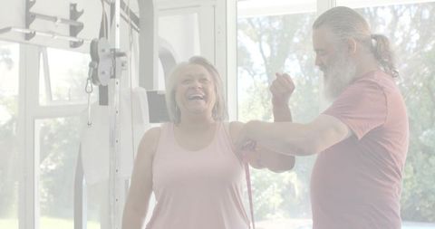 Senior woman laughing during assisted resistance band exercise with trainer in bright gym