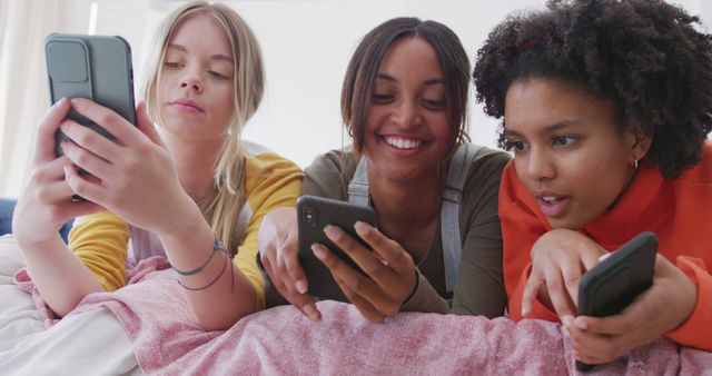 Happy diverse teenager girls lying on bed and using smartphones in bedroom. Spending quality time, lifestyle, friendship and adolescence concept.