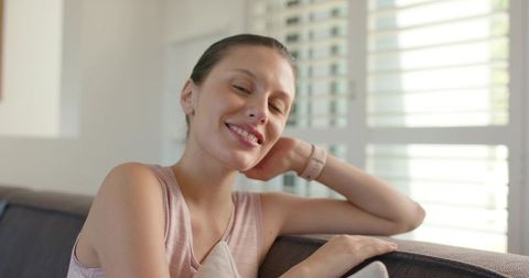 Smiling young woman relaxing on couch with sunlit shutters pastel minimalist cozy interior