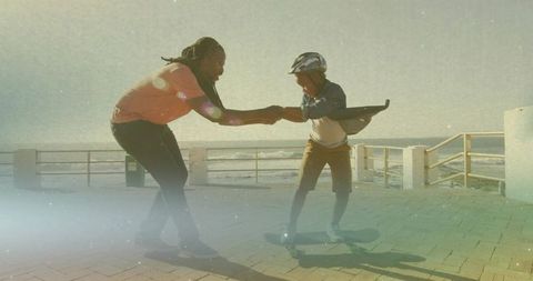 Man teaching child skateboarding on coastal boardwalk hand-holding helmeted safety guidance