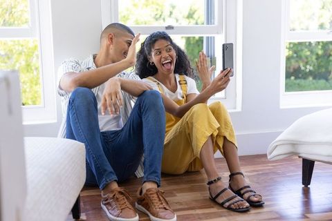 Couple celebrating on hardwood floor while video calling in bright modern home interior