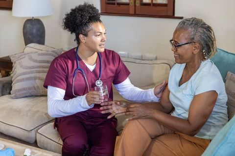 Home nurse offering water and comforting senior woman on sofa during in-home care visit