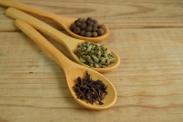 Close-up shows three wooden spoons filled with different spices including allspice, herbs, and cloves, placed on a wooden surface. This image is perfect for blogs, articles, and recipes focused on cooking, seasoning techniques, culinary arts, or natural ingredients. Use it in promotional materials for food products or culinary schools.