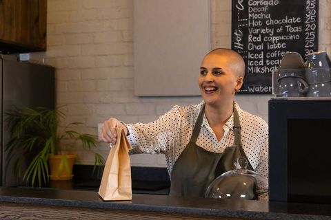 Smiling barista handing takeaway bag at cafe counter wearing gray apron, polka-dot shirt