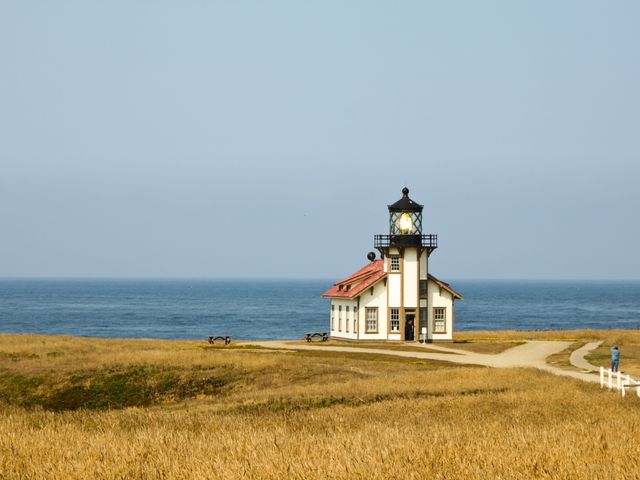 Scene shows a solitary lighthouse beside the ocean on a clear, sunny day. The lighthouse stands in a grassy field with a path leading up to it. This image can be used for travel blogs, maritime publications, tourism promotions, or coastal-themed designs.