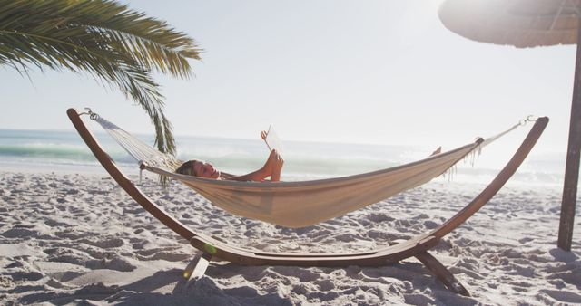 Woman lying in hammock on sandy beach under palm trees, enjoying vacation and reading book by the ocean. Suitable for travel websites, vacation promotions, relaxation concepts, tropical destination advertisements, or leisure lifestyle blogs.