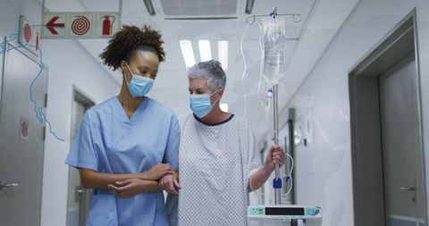 Nurse helping senior patient walk with iv drip in hospital corridor wearing masks
