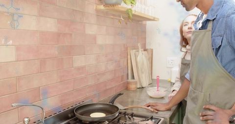 Diverse couple cooking pancakes on gas stove with pink tile backsplash and cozy kitchen