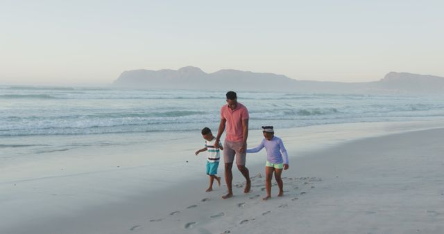 Father walks hand in hand with two children along a sandy beach at dawn. Perfect for conveying themes of family bonding, relaxation, and travel. Useful for advertisements promoting family vacations, beachfront resorts, and lifestyle content focused on family values and quality time with loved ones.