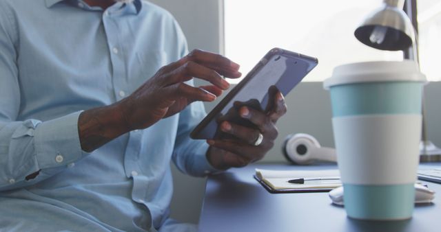 The image depicts a professional man sitting at a desk in an office environment, using a tablet. The setting is modern and casual, suggesting a contemporary work lifestyle. The focus on his hands emphasizes engagement and productivity. The scene includes a coffee cup and a desk lamp, adding to the professional ambiance. This image can be used in articles or marketing materials related to business, modern work environments, technology use, productivity, and remote working setups.