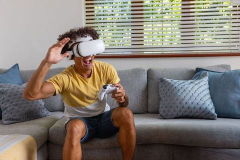 Young man playing vr game on couch reacting with joy while wearing headset and holding controller