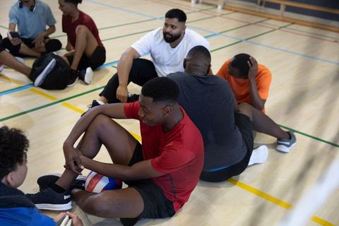 Multicultural male teammates sitting on gym court holding red-white-blue ball and chatting