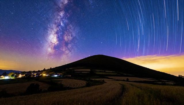 Milky way rising with star trails over rolling hill and village lights