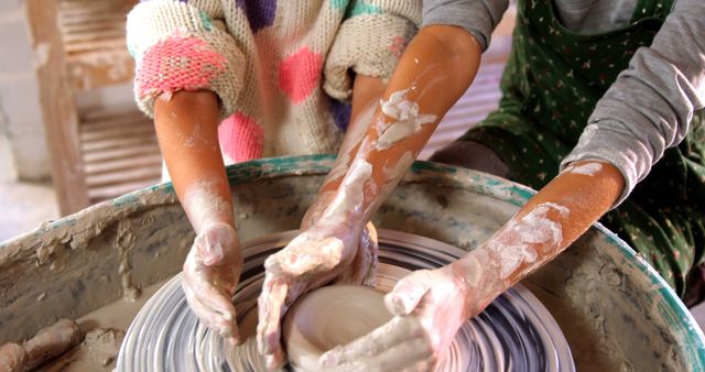 This image shows a close-up of two pairs of hands crafting pottery on a pottery wheel, highlighting the collaborative effort in creating ceramic art. Perfect for use in websites or marketing materials focused on arts and crafts, pottery classes, artisan workshops, and creativity promotion.