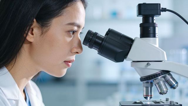 Asian researcher examining slide through microscope in clinical lab close-up precision focus