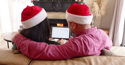 Couple wearing santa hats sharing tablet while watching fireplace on cozy sofa