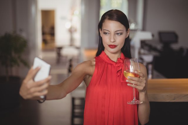 Woman in red dress taking selfie with mobile phone while holding glass of wine in restaurant. Ideal for use in lifestyle blogs, social media content, advertisements for restaurants or wine brands, and articles about modern dining experiences.