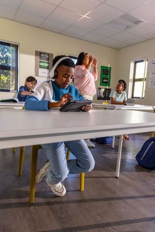 Boy wearing headset using tablet in classroom while teacher guiding diverse students