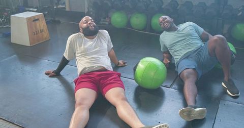 Male friends resting on gym floor after workout, leaning back with green medicine ball