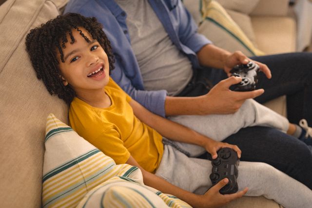 Portrait of hispanic boy playing video games with father sitting on sofa in living room at home. unaltered, technology, enjoyment, family, lifestyle and togetherness concept.