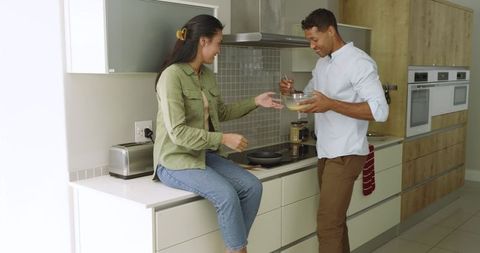 Smiling diverse couple cooking and laughing in sunlit kitchen sharing mixing bowl breakfast