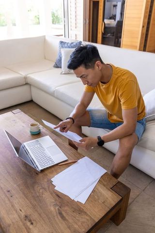 Asian man sitting on sofa reviewing documents with laptop in sunlit living room