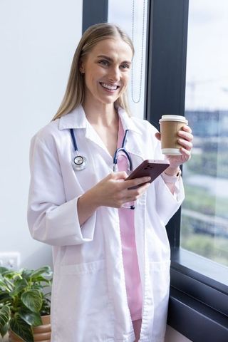 Smiling female doctor holding phone and coffee wearing white coat and pink scrubs by window