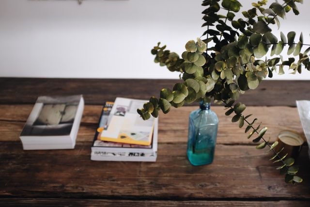 Vintage wooden table decorated with a blue vase holding eucalyptus branches. Several stacks of books and magazines lie next to the vase. Ideal for themes of rustic decor, minimalist interior design, home workspace inspiration, and relaxation.