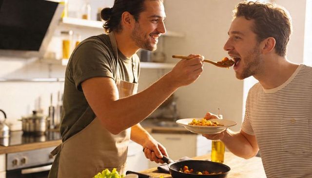 Sunlit kitchen couple cooking together, feeding each other pasta and sharing smiles