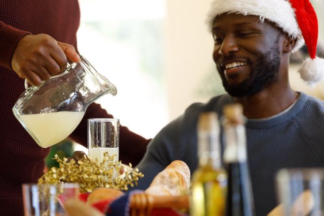 African-American man wearing a Santa hat, smiling while sitting at a Christmas dinner table. Another person is pouring a drink into a glass. The table is set with festive decorations and food items. Ideal for use in holiday-themed promotions, advertisements, and social media posts celebrating Christmas and family gatherings.