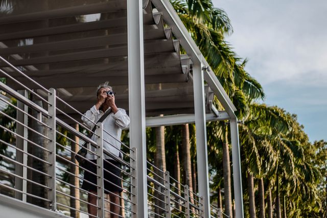 This image shows a man on a balcony capturing photographs with a camera, surrounded by tropical palm trees. Ideal for illustrating travel blogs, vacation advertisements, photography themes, or lifestyle articles focused on outdoor activities and sightseeing.