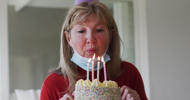 A woman is blowing out birthday candles on a cake while wearing a mask under her chin. She also wears a party hat, implying a birthday celebration during the COVID-19 pandemic era. This image can be used for articles about pandemic celebrations, personal milestones, or birthday wishes during COVID-19.