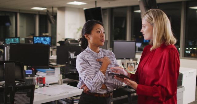Two businesswomen engaged in discussion in a modern office environment at night, with desktop computers and office equipment in the background. Perfect for representing teamwork, female professionals in business, corporate environment, night shifts, and collaborative workspaces. Useful for articles, blogs, marketing materials, and advertisements related to business, productivity, women in the workforce, and professional development.