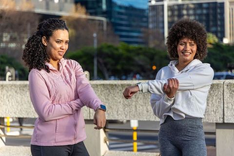 Diverse women stretching arms on urban waterfront before outdoor workout wearing smartwatches