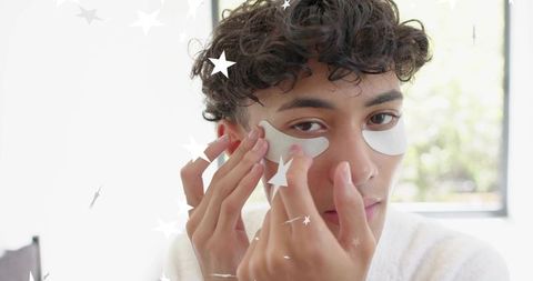 Adult male applying hydrogel under-eye patches wearing white robe near sunlit window
