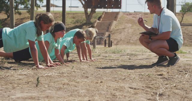 Group of children wearing matching teal shirts performing push-ups under the guidance of a coach outdoors. Might be great for illustrating themes such as youth sports camps, physical fitness, teamwork, outdoor activities, and healthy living for kids. Suitable for use on websites and marketing materials promoting youth athletics, sports training programs, or summer camps.