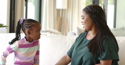 African american mother and daughter sitting and smiling on sofa in bright living room